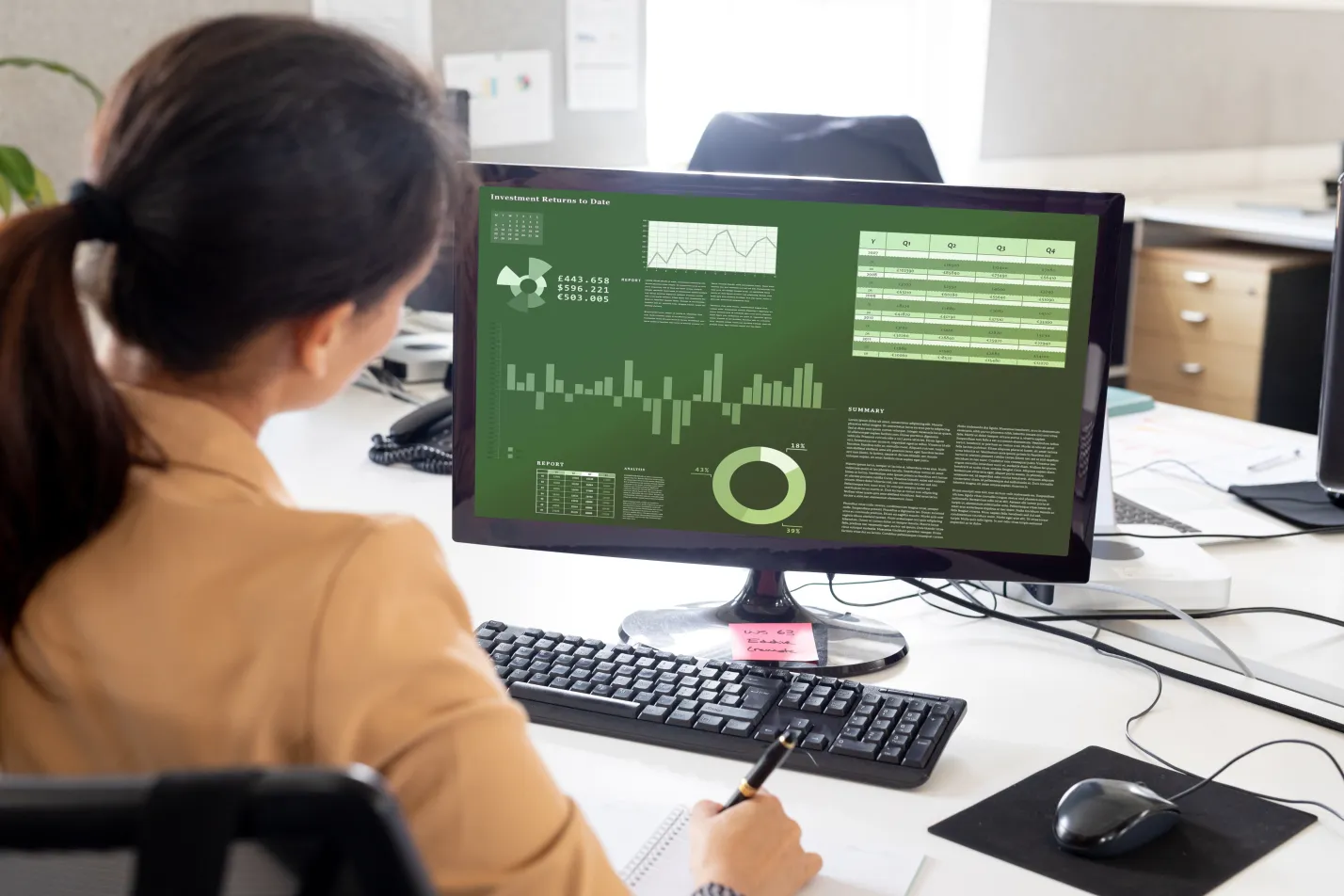 Woman at a desk taking notes while reviewing a data dashboard on her monitor
